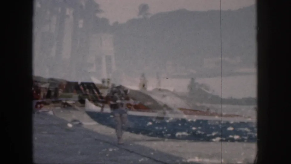 A soft 1959 Hawaii waterfront frame with boats, bright water reflections, and a walking figure partly blurred by motion.