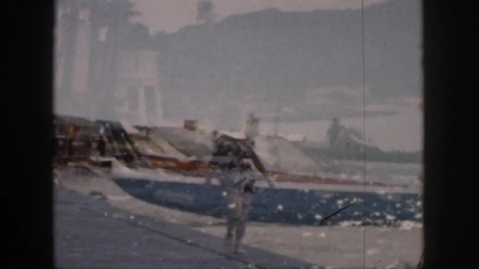 1959 Hawaii home-movie frame showing a blurred man near docked boats on a waterfront with a hazy shoreline beyond.