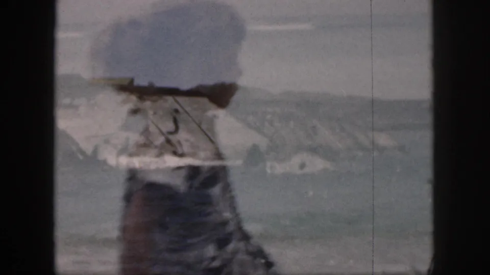 1959 Hawaii home-movie frame with surf, distant hills, and a beachgoer in a patterned dress partly obscuring the foreground.