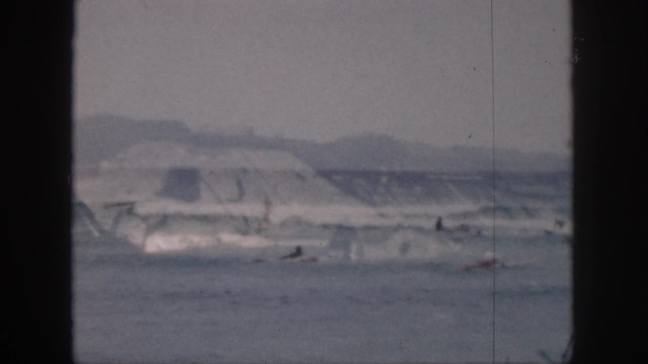 Distant surfers ride low breaking waves beneath hills and sky.