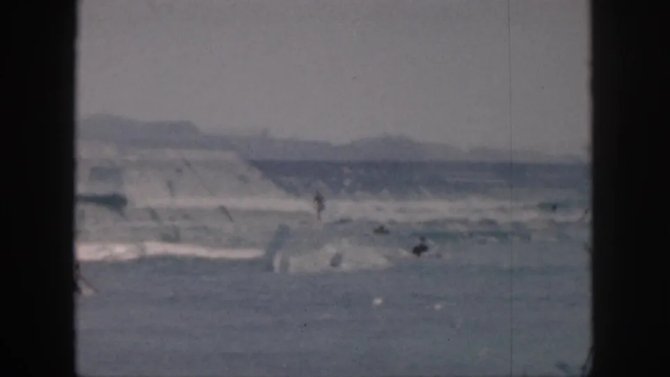 Faded 8mm Hawaii beach frame from 1959 showing scattered surfers or swimmers in rough surf below distant hills.