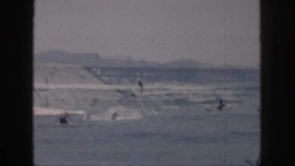 1959 Hawaii 8mm frame with choppy surf, distant hills, and small beachgoers or surfers spread across the water.