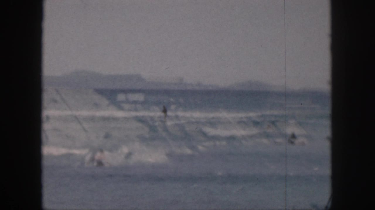 Distant surfers and swimmers in small waves, framed by dark film borders.