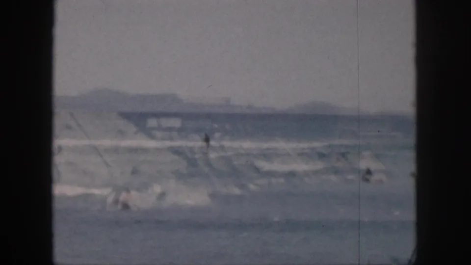 1959 Hawaii home-movie frame showing distant people in the surf, soft shoreline shapes, and dark 8mm frame borders.
