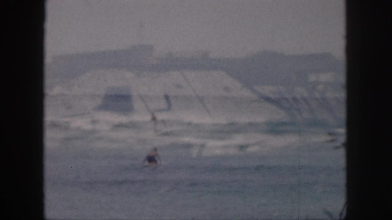 A distant surfer or swimmer sits in the water beyond low breaking waves.