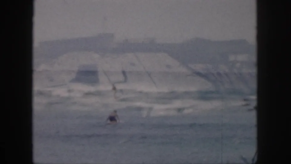 1959 Hawaii surf scene with one distant figure in the water, pale waves, hazy hills, and soft home-movie focus.