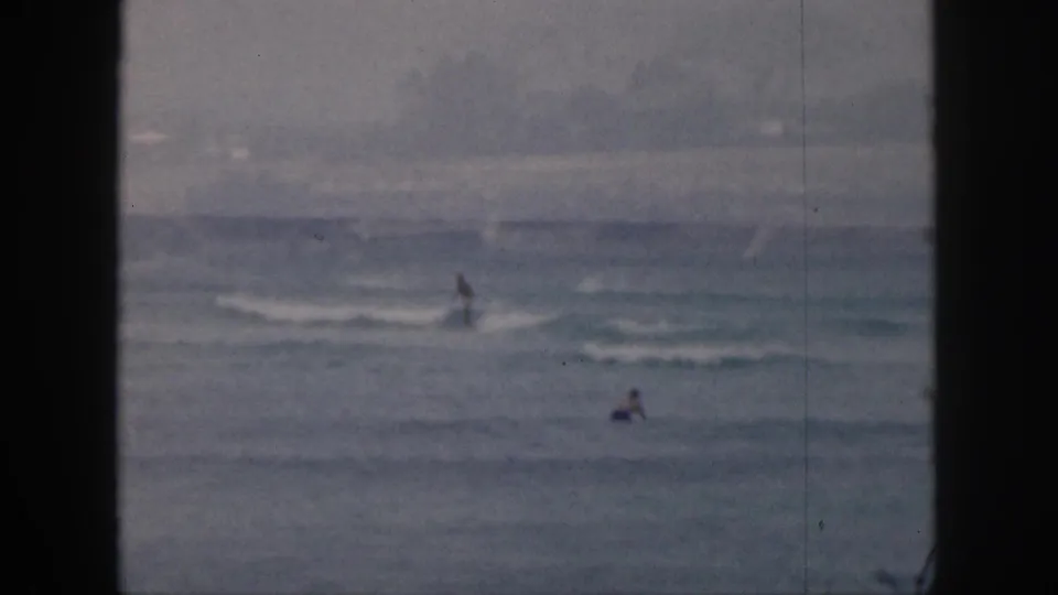 1959 Hawaii beach footage frame with distant surfers or swimmers, rolling small waves, and muted 8mm color.