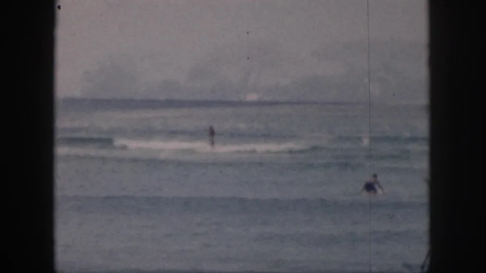 1959 Hawaii home-movie frame showing distant bathers or surfers in gentle waves beneath an overcast sky.