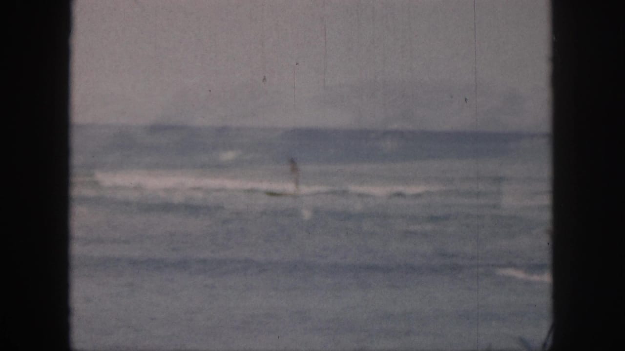 1959 Hawaii 8mm frame with a distant lone surfer moving through gentle surf, framed by dark film borders.