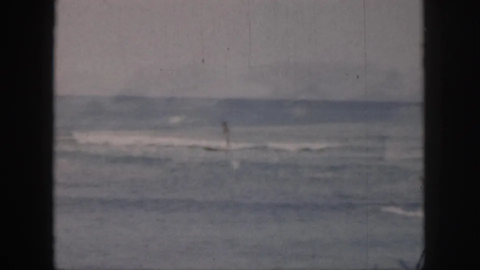 1959 Hawaii 8mm frame with a distant lone surfer moving through gentle surf, framed by dark film borders.