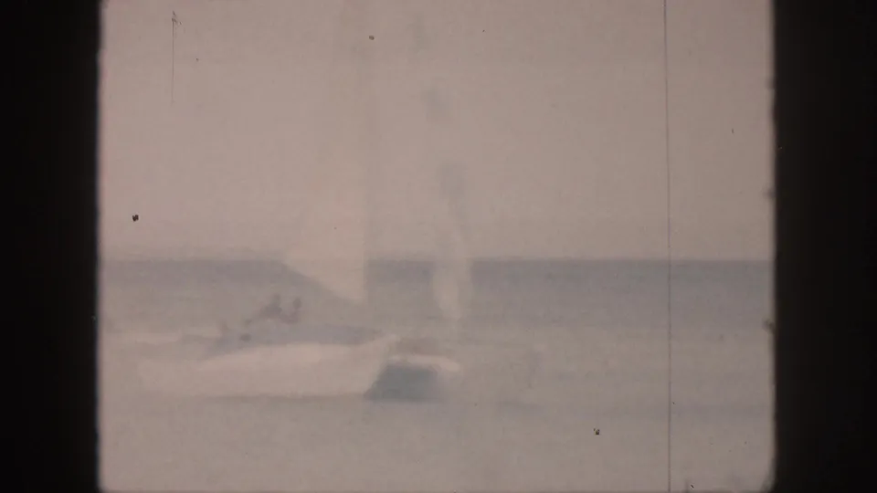 1959 Hawaii 8mm frame showing a white sailboat on calm ocean water with small figures aboard and a hazy horizon.