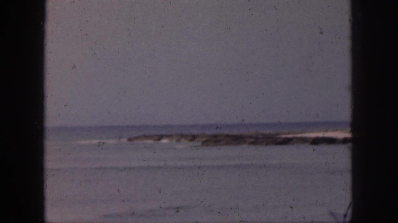 1959 Hawaii coastal view with calm water, a hazy horizon, and a distant reef or rocky shoreline behind dark framing.