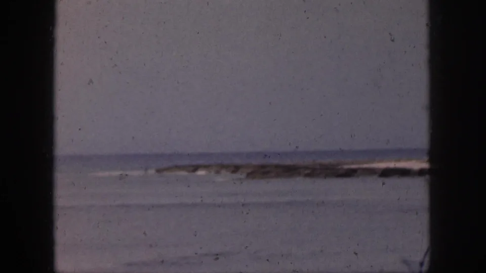 1959 Hawaii coastal view with calm water, a hazy horizon, and a distant reef or rocky shoreline behind dark framing.