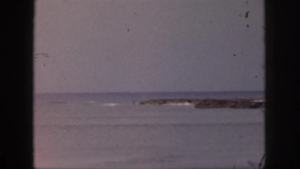 1959 Hawaii shoreline footage showing muted water, low waves, and a distant rocky or reef line under overcast haze.
