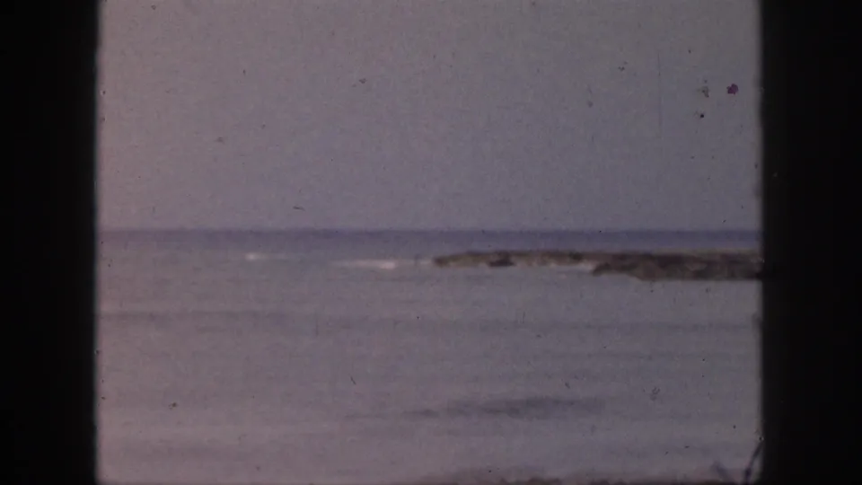 1959 Hawaii home-movie still of calm coastal water, a distant reef or shoreline, and dark window-frame edges.