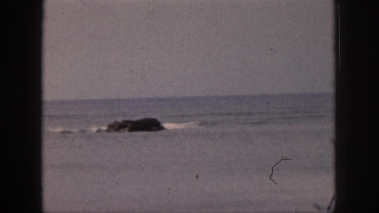 1959 Hawaii 8mm frame showing a dark rock in calm ocean water, with soft sky and visible film dust through a window-like frame.