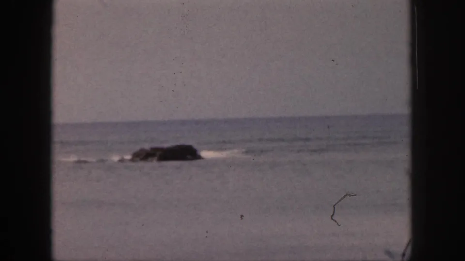 1959 Hawaii 8mm frame showing a dark rock in calm ocean water, with soft sky and visible film dust through a window-like frame.
