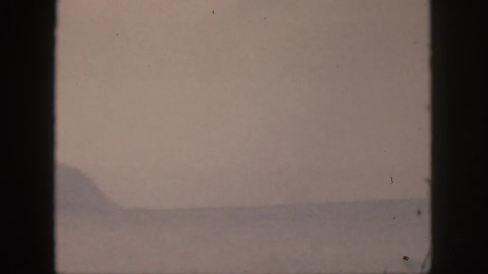 1959 Hawaii home-movie frame with a distant coastal landform across calm water, softened by grain, haze, and muted exposure.
