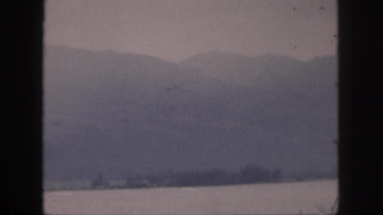 1959 Hawaii home-movie view of hazy mountains and shoreline across water, with a dark window bar in the foreground.