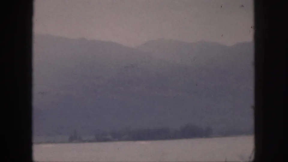 1959 Hawaii home-movie view of hazy mountains and shoreline across water, with a dark window bar in the foreground.