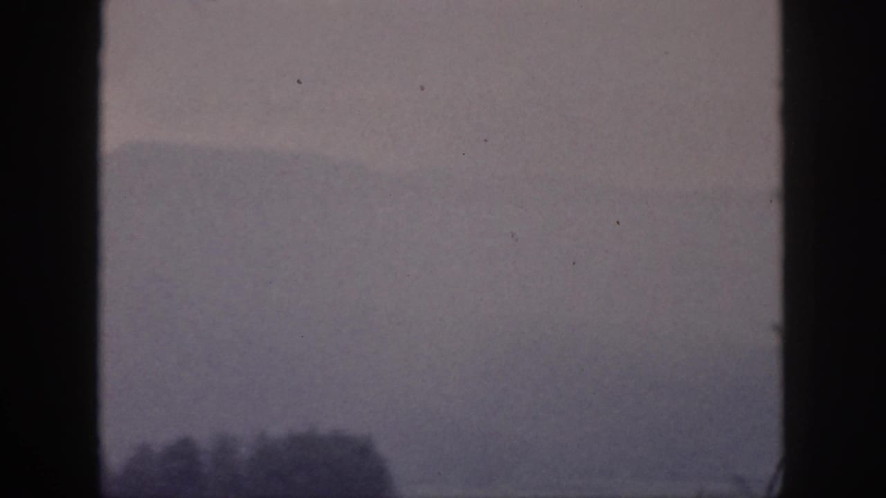 1959 Hawaii home-movie frame with a pale mountain ridge, mist, and dark tree shapes beneath vertical window framing.