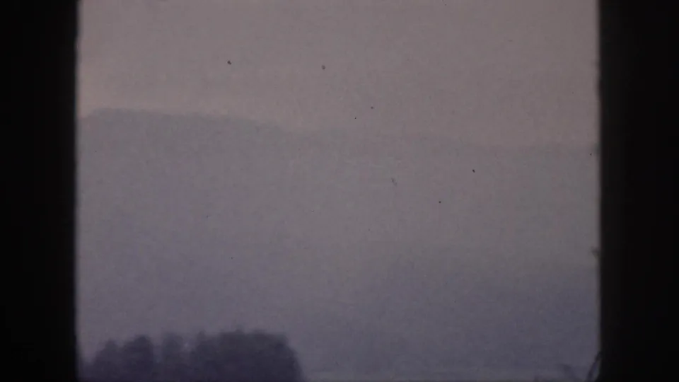 1959 Hawaii home-movie frame with a pale mountain ridge, mist, and dark tree shapes beneath vertical window framing.