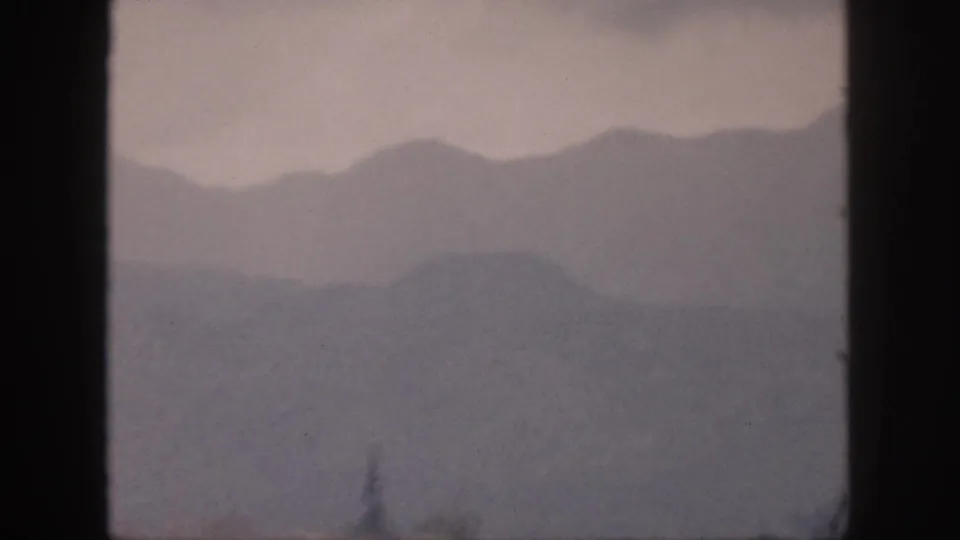 1959 Hawaii home-movie frame showing muted mountain silhouettes through a window, with faint vegetation near the lower edge.