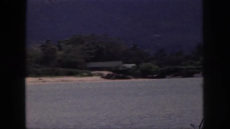 1959 Hawaii 8mm coastal view with calm ocean water, a sandy strip, dense shrubs, and a hazy mountain behind.