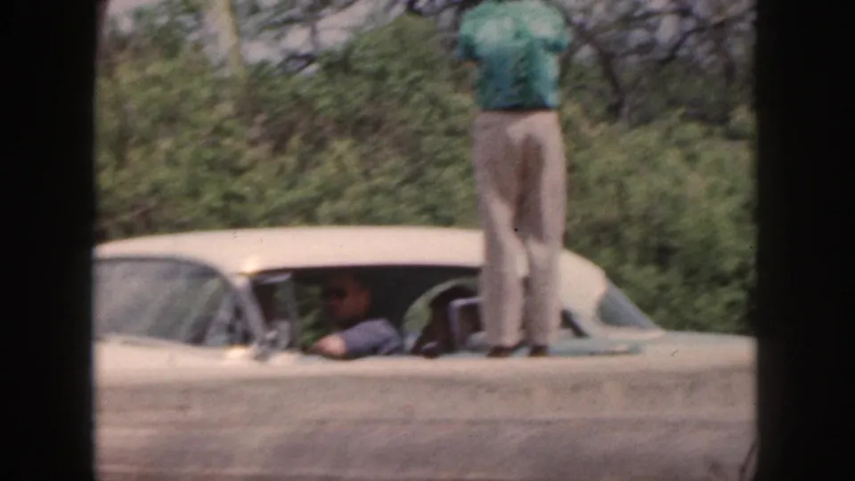 1959 Hawaii home-movie frame with a man in a green shirt standing on a white car roof as another person sits in the driver area.