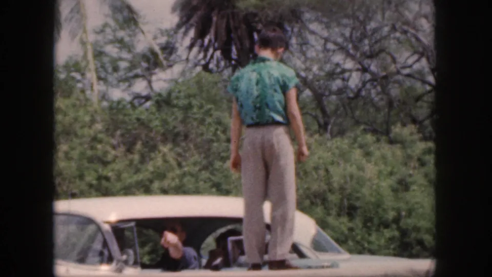 1959 Hawaii home-movie frame showing the rear of a man standing on a white car roof, with another person partly visible inside.