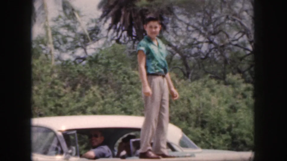 1959 Hawaii home-movie frame with a man on a white car roof turning toward the camera near tropical roadside trees.