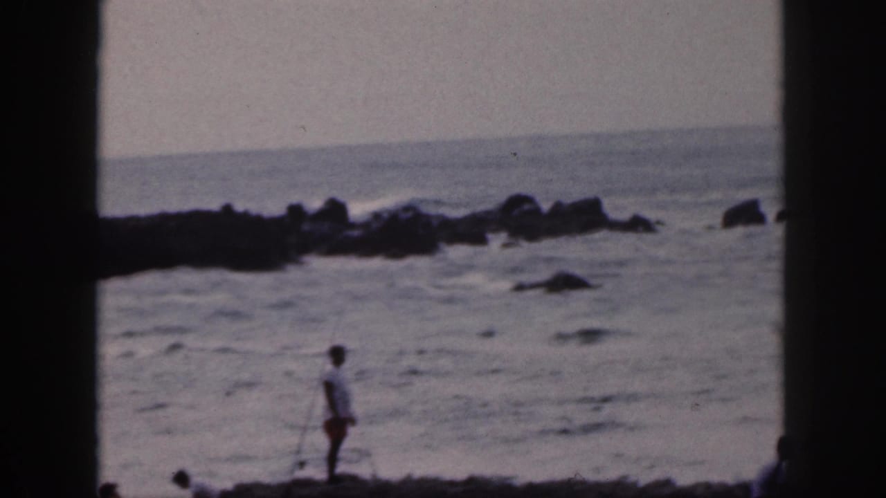 1959 Hawaii 8mm frame of a person in a white shirt standing with a fishing pole beside a rocky, windblown shoreline.
