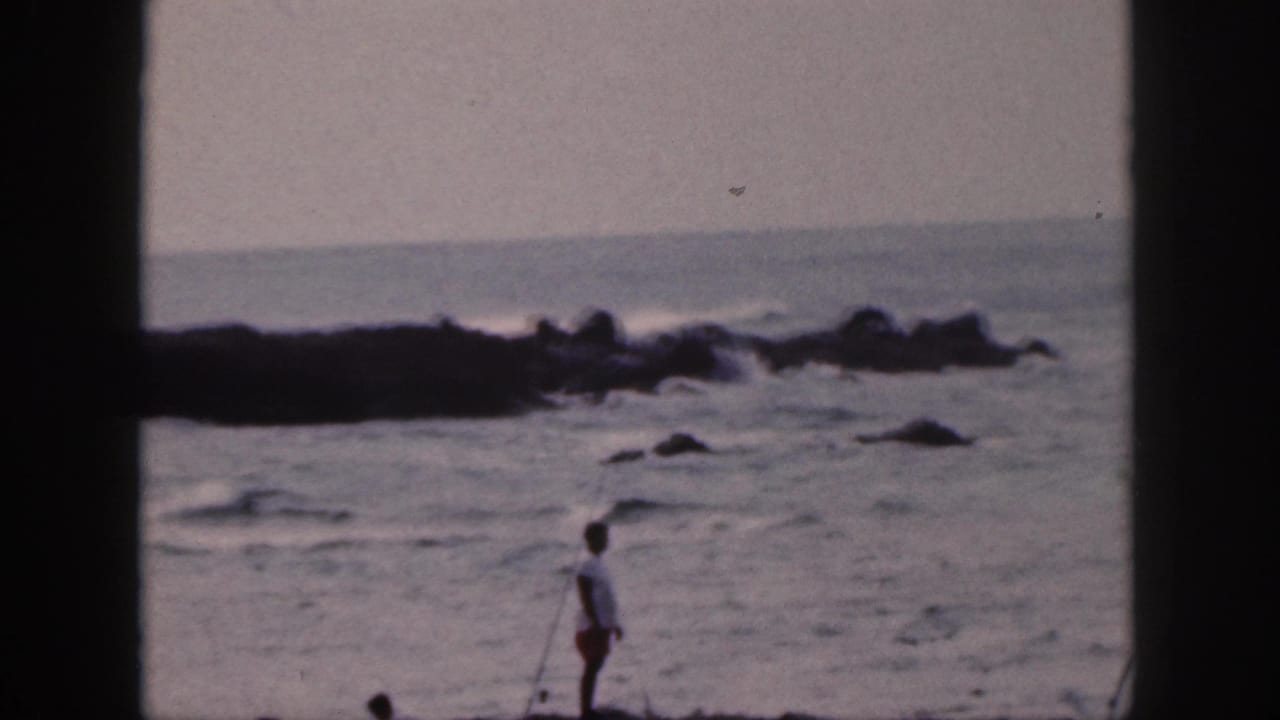 Fisher stands at the water's edge with surf and rocks behind.