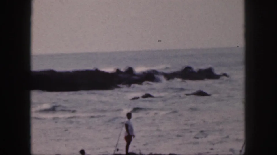 1959 Hawaii home-movie frame showing a distant fisher at the edge of rough water and dark coastal rocks.