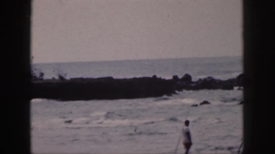 1959 Hawaii 8mm frame with muted surf, dark rocks, and a distant person partly low in the home-movie composition.