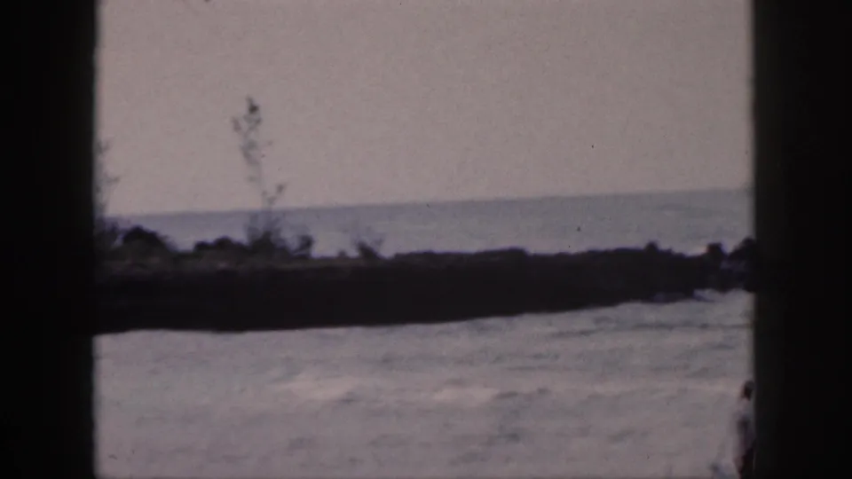 1959 Hawaii home-movie frame of surf washing past dark shoreline rocks, with the fisher partly cropped at the right edge.