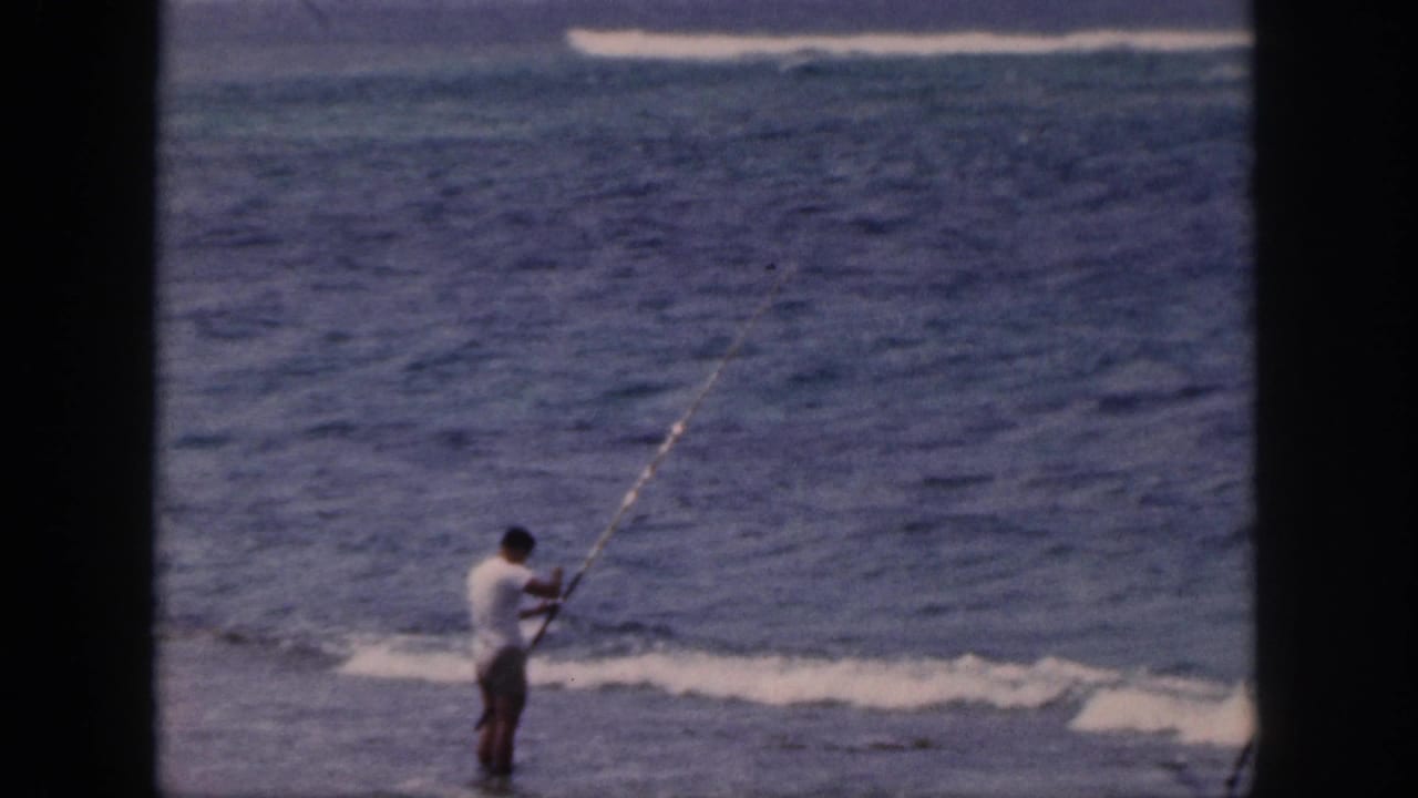 1959 Hawaii home-movie frame showing a man in a white shirt fishing from the shoreline beside blue ocean water.