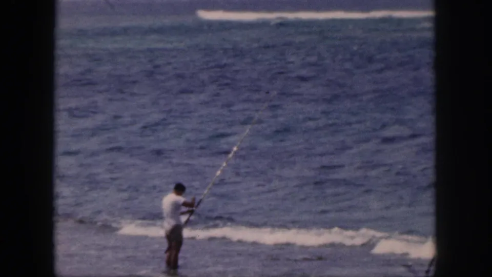 1959 Hawaii home-movie frame showing a man in a white shirt fishing from the shoreline beside blue ocean water.