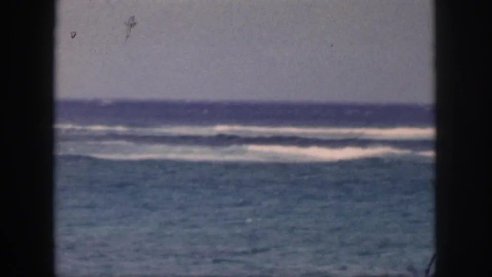 Archival 8mm view of Hawaii surf, with turquoise water, distant white breakers, and dark vertical framing at the sides.
