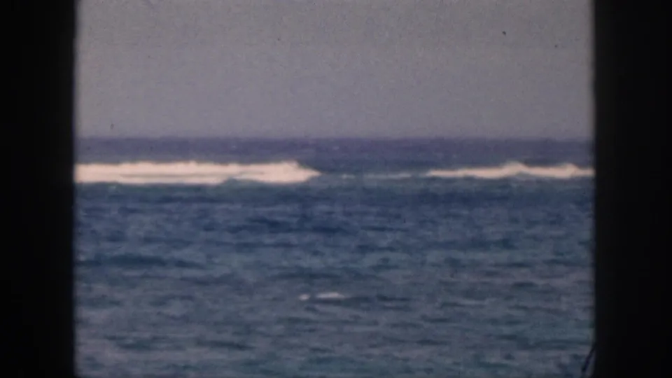 Archival Hawaii surf view with blue water, white foam, distant horizon, and a dark doorway or window edge in the foreground.