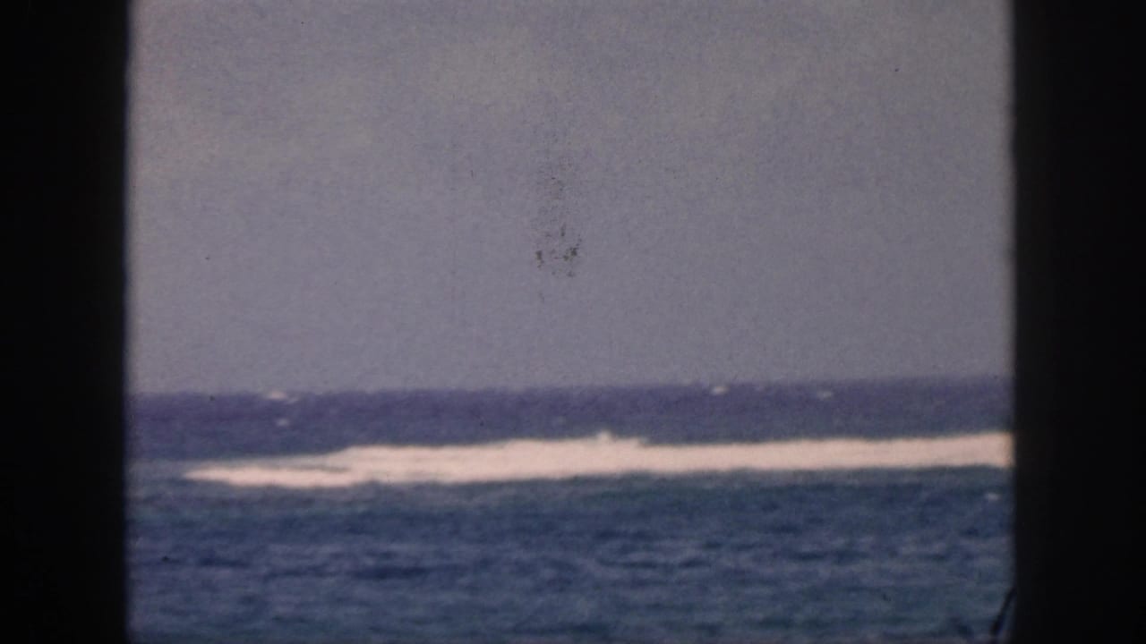 1959 Hawaii home-movie frame showing blue ocean, distant white surf, gray sky, and dark window bars in the foreground.