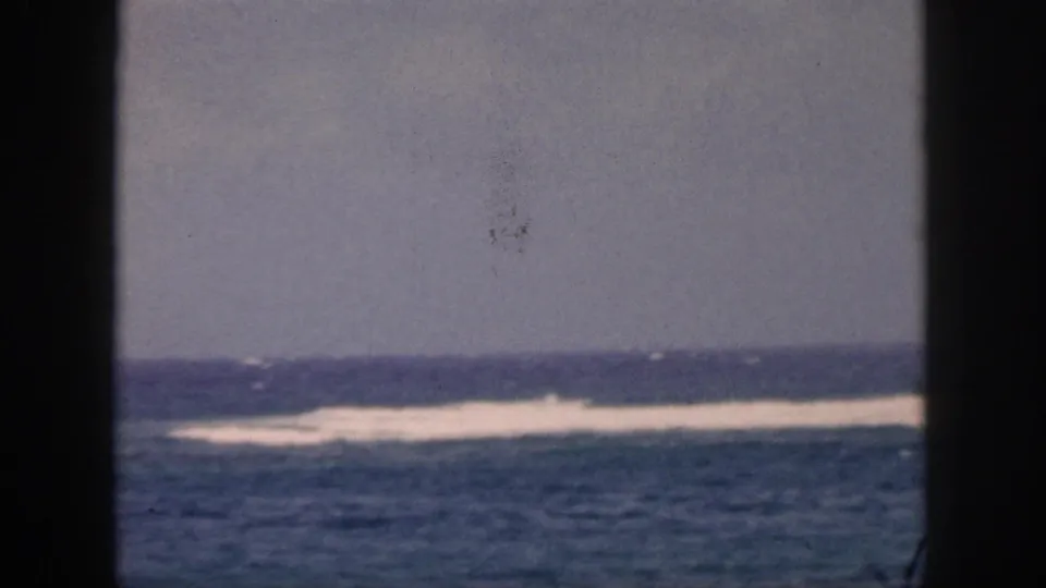 1959 Hawaii home-movie frame showing blue ocean, distant white surf, gray sky, and dark window bars in the foreground.