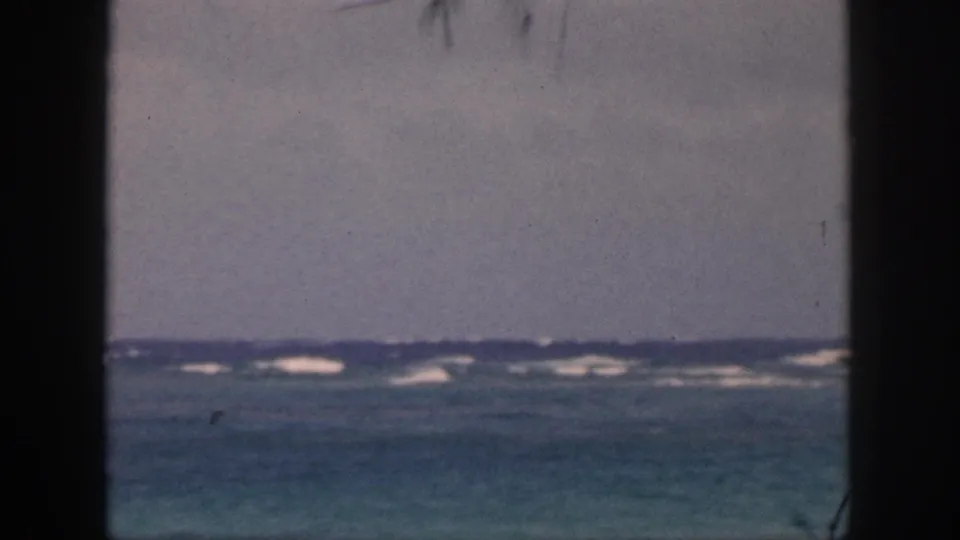 1959 Hawaii 8mm frame showing low ocean horizon, white breaking waves, pale sky, and dark vertical framing at the sides.