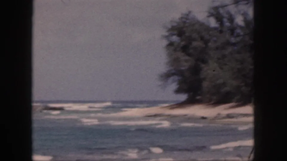 1959 Hawaii 8mm frame showing a sandy coastal point with dense trees and surf, partly blocked by dark window framing.