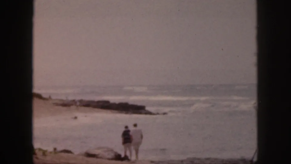 1959 Hawaii 8mm frame with a couple walking along the beach beside gentle surf and a low rocky edge.