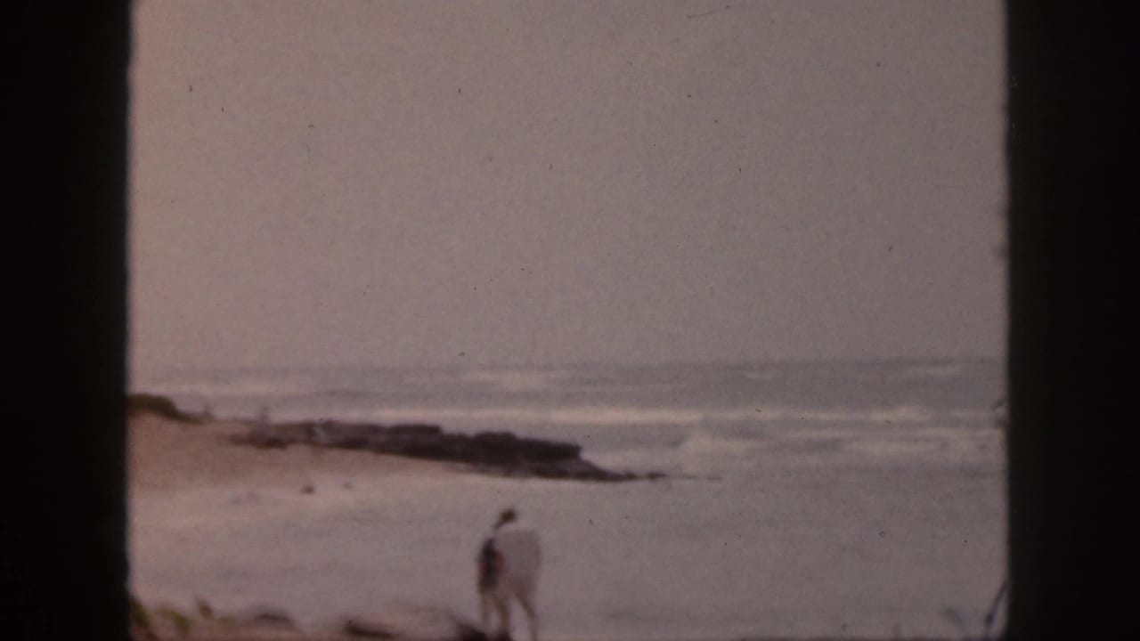 1959 Hawaii 8mm frame showing two distant adults near a rocky shoreline under a gray sky.