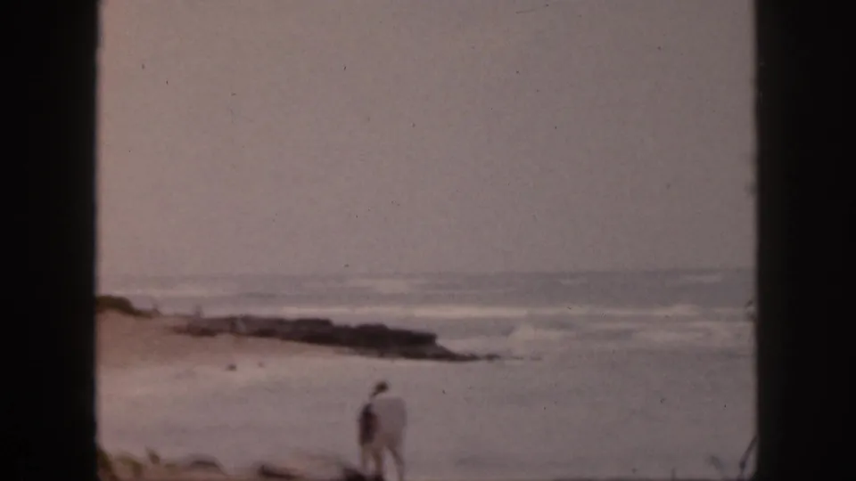 1959 Hawaii 8mm frame showing two distant adults near a rocky shoreline under a gray sky.