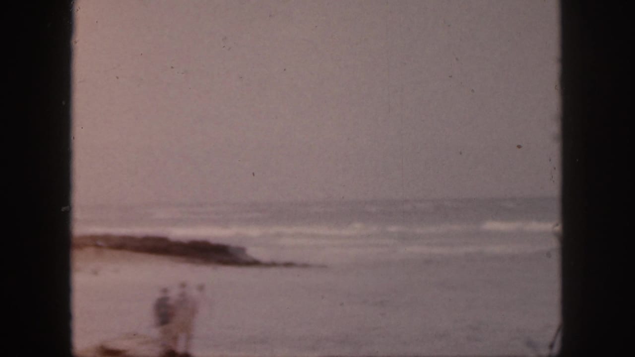 Blurred figures stand or walk near the shoreline with rocks and ocean behind them.