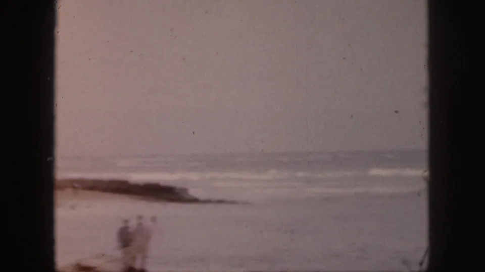 Soft 8mm Hawaii beach view with two distant adults near surf and dark rocks.
