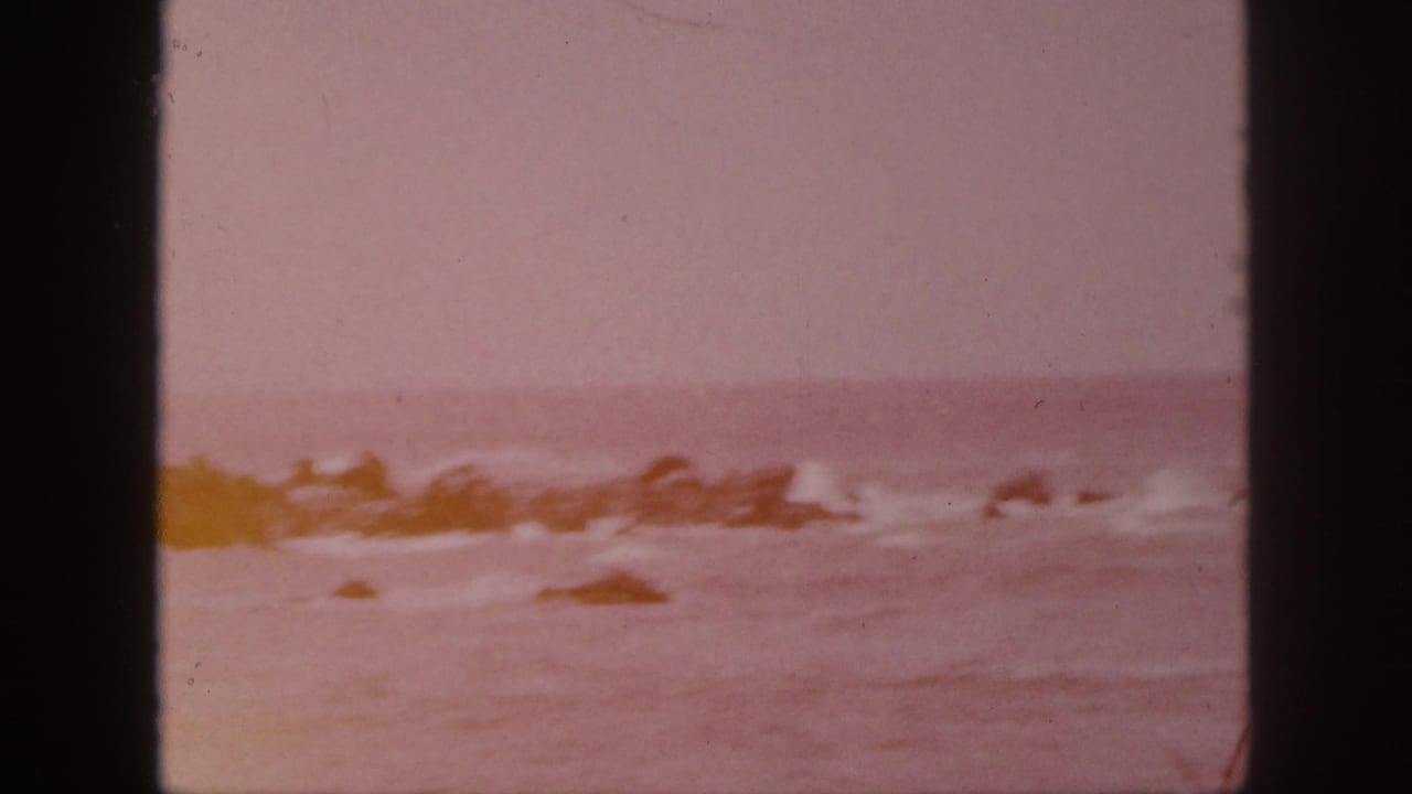 1959 Hawaii 8mm frame showing surf breaking around dark rocks beneath a pale sky, with faded color and black film borders.